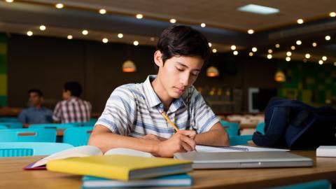 Young boy sitting at a desk with books, a computer, and a pencil he is using to write in a notebook.