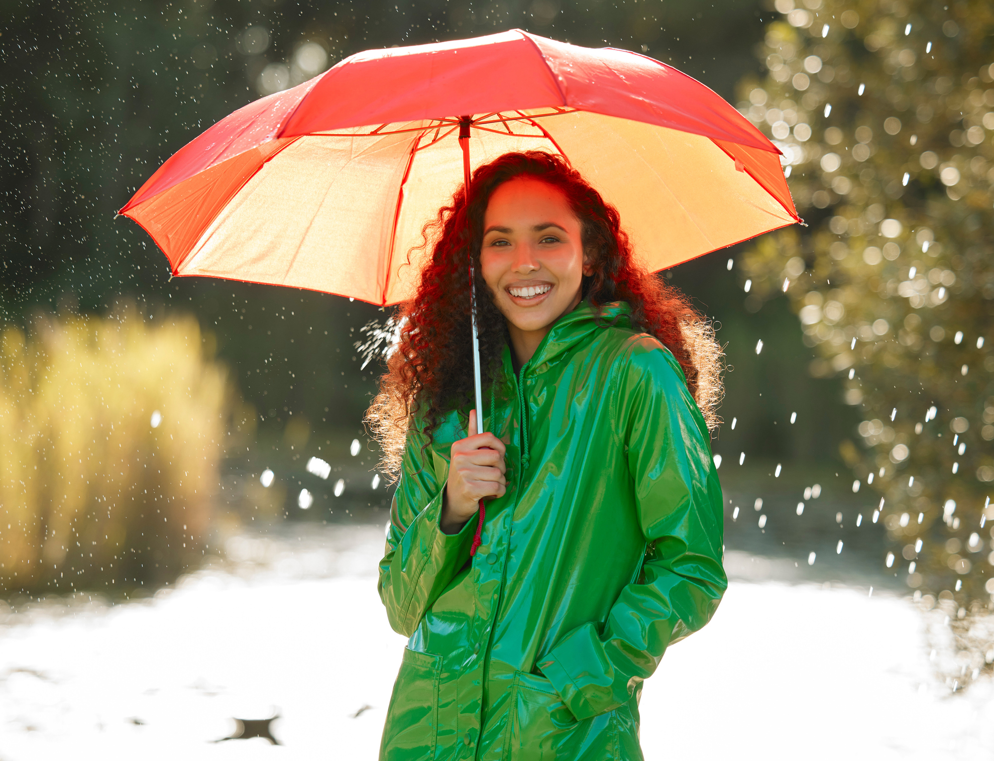 Woman in a green rain jacket standing in the rain holding a red umbrella