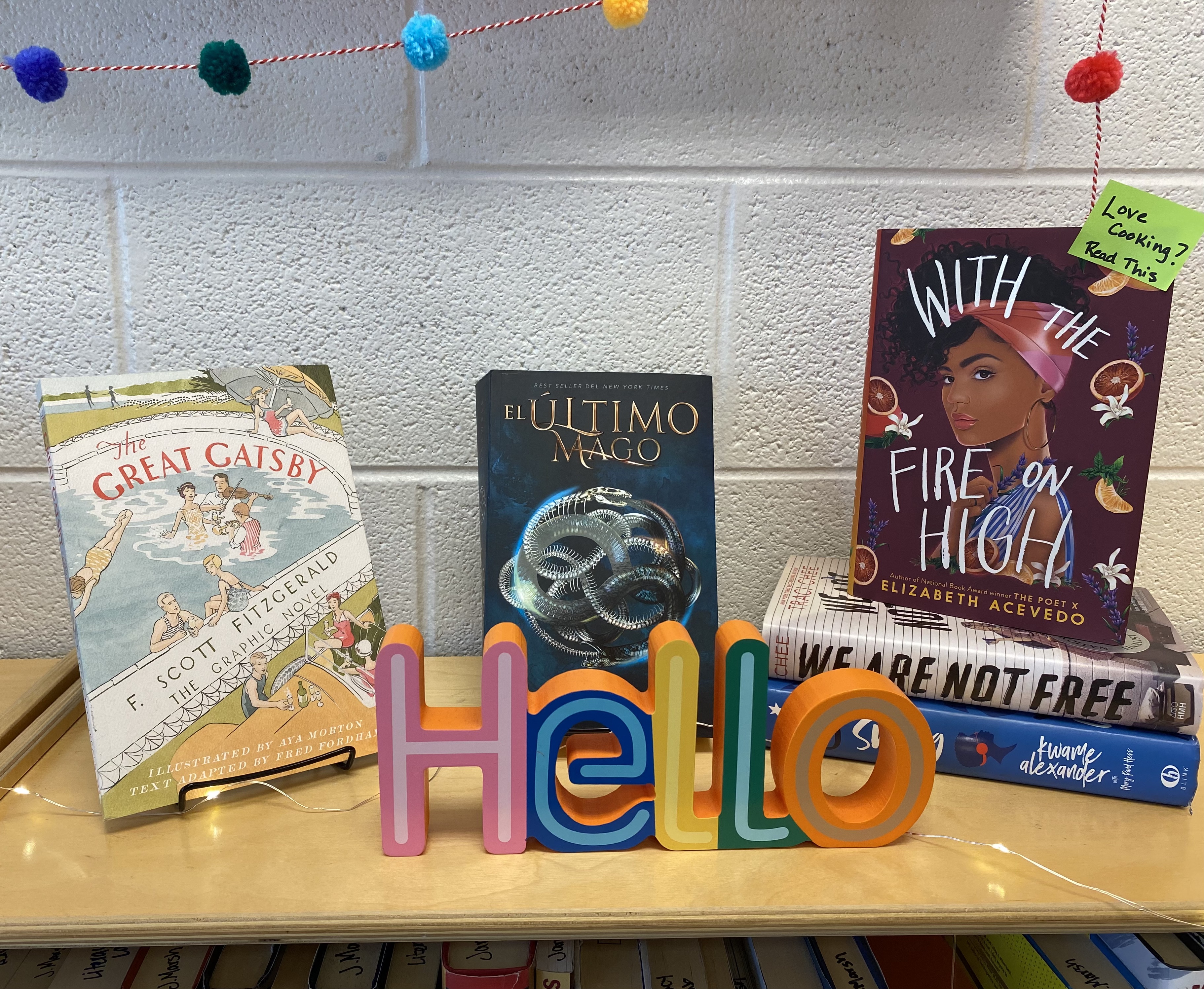 YA BOOKS stacked on a low bookshelf in a teacher's room with a 'hello' sign in front.