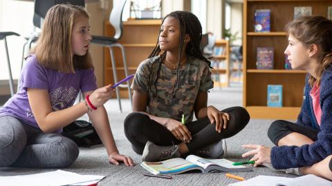 Three middle school girls sitting on floor of school library talking about schoolwork