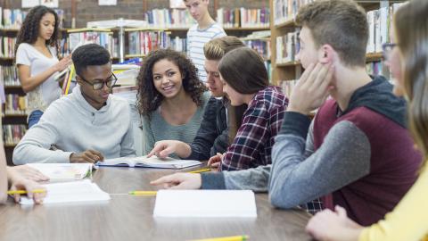 A diverse group of teenagers sitting at a library table discussing a text in front of them.