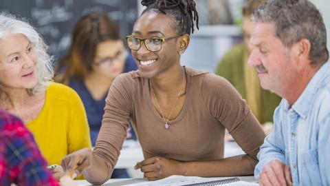 A small group of mature adults sit together as they work collectively on a group project. They are each dressed casually and have notebooks and papers out in front of them as they reference the material for the project.