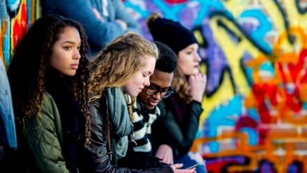 Multicultural group of four teens outside in front of colorful mural with two looking at cell phone