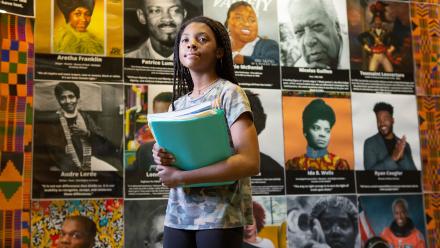 Black middle school girl holding notebooks in front of display of Black history icons