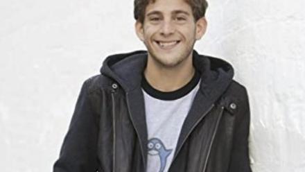 Adi with a black leather jacket leaning on a white wall