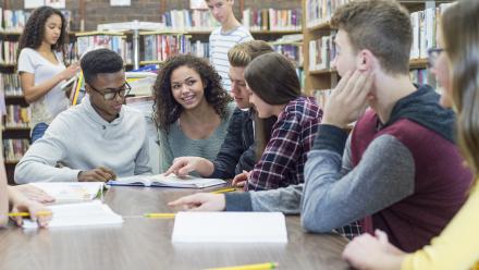 A diverse group of teenagers sitting at a library table discussing a text in front of them.