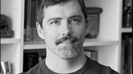 black and white picture of the author sitting in front of a bookcase behind him