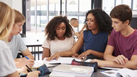 High school students at a table with a teacher pouring over reading material