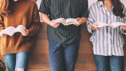 Bodies of three teens holding books, standing against a low, wood wall