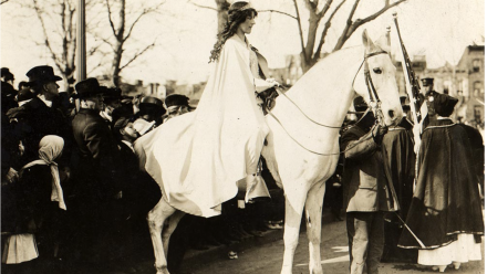 1920's woman sitting on a white horse while wearing a long white cape and tiara.