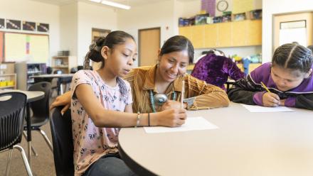 A teacher is seated between two ELL students working on classwork
