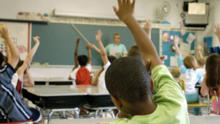 Different races of students seated in a classroom, seated and raising their hands to be called on by the teacher