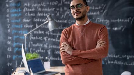 young, male teacher leaning against his desk with chalk writings on the board behind him
