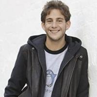 Adi with a black leather jacket leaning on a white wall 