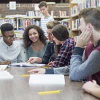 A diverse group of teenagers sitting at a library table discussing a text in front of them.