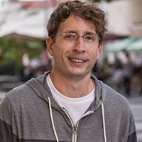 Headshot of Shaun wearing a grey hooded sweatshirt and glasses