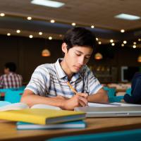 Young boy sitting at a desk with books, a computer, and a pencil he is using to write in a notebook.