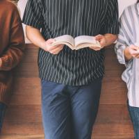 Bodies of three teens holding books, standing against a low, wood wall