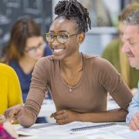 A small group of mature adults sit together as they work collectively on a group project. They are each dressed casually and have notebooks and papers out in front of them as they reference the material for the project.