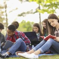 Group of teenagers reading books while sitting on the ground outside.